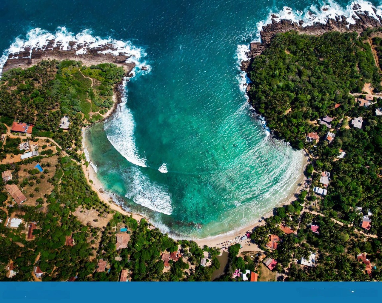 Aerial view of a Sri Lankan bay with turquoise waters, white surf, lush tropical greenery and red-roofed houses